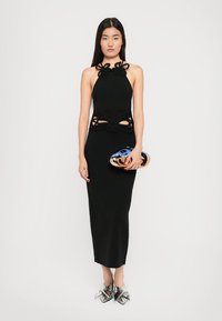 Rochie neagră cu gât halter, cu decupaje la talie, siluetă lungă și țesătură texturată. Modelul ține în mână o poșetă metalică și poartă pantofi argintii.
