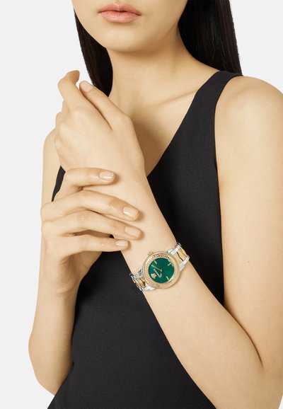 Metallkellaga kellal on roheline sihver, kullast ja hõbedast roostevabast terasest rihm, ümmargune nägu dekoratiivse äärisega ja õhukesed käed.