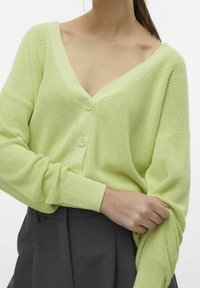 Cárdigan de punto verde claro con escote en V, con dos botones, puños acanalados y mangas holgadas. Lleva puesto sobre pantalones de talle alto grises.
