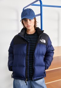 Marineblå puffjakke med sorte skulderpaneler, glidelås og lommer. Kombinert med marineblåstripet skjorte og denimcaps.