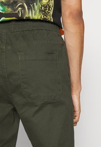 Olijfgroene broek met een achterzak voorzien van een rechthoekige klep en zichtbare stiksels, gemaakt van een lichtgewicht katoenmix.