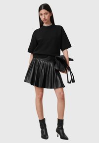 Maglione nero oversize con maniche corte, abbinato a una gonna in pelle plissettata nera. La modella tiene una borsa nera e indossa stivali alla caviglia neri.