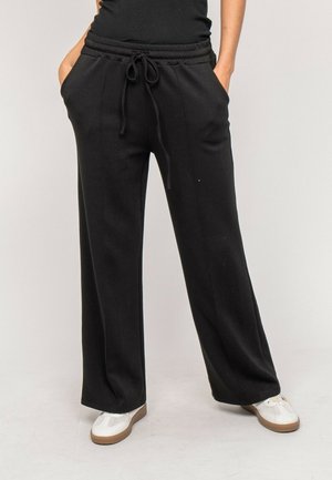 Personne portant un pantalon noir à jambes larges avec taille à cordon et des baskets blanches, les mains dans les poches, debout sur un sol clair.