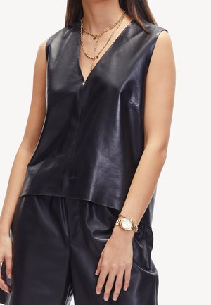 Femme portant un haut en cuir noir sans manches à col en V et un pantalon assorti, accessoirisée avec des colliers superposés en or et une montre en or.