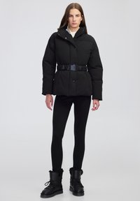 Donna in piedi che indossa un piumino nero con cintura, leggings neri e stivali neri in pelle foderati di pelliccia con lacci, su uno sfondo chiaro e semplice.