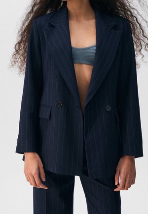 Vrouw draagt een donker gestreept colbert over een geribde cropped top, met bijpassende gestreepte broek, staand tegen een effen achtergrond.