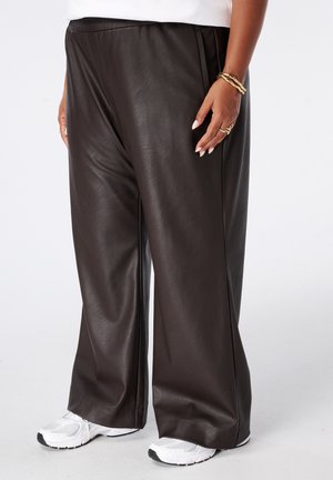 Persoon draagt losse donkerbruine leren broek, witte sneakers, witte top en gouden ringen en armbanden aan de rechterhand.