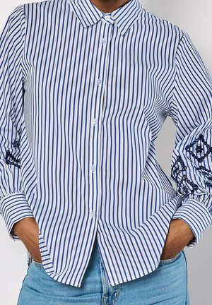 Kobieta w koszuli na guziki w pionowe, niebiesko-białe paski z ciemnoniebieskimi haftowanymi wzorami na rękawach oraz w jasnoniebieskich dżinsach.