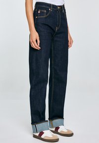 Jeans de mezclilla azul oscuro con un diseño de pierna recta, que presentan costuras amarillas, bolsillos frontales y puños doblados que muestran un detalle de mezclilla más clara.
