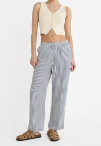Vrouw in een crème kleurige mouwloze button-up crop top, blauwe en witte verticaal gestreepte wijde broek en tan kleurige gevlochten sandalen.
