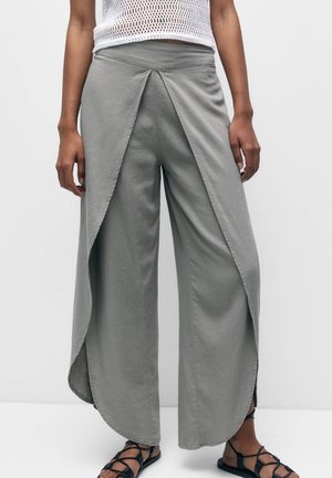 Woman wearing light gray wide-leg pants with wrap overlay and a white sleeveless knit top, paired with black strappy sandals.