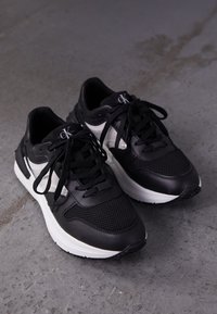 Černé sportovní tenisky s vrškem z prodyšné síťoviny a kůže, bílou podrážkou, kulatou špičkou, černými tkaničkami a logem na jazyku.