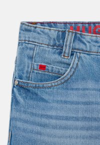 Jeans in denim azzurro con un'etichetta rossa "HUGO". Caratteristiche: cuciture arancioni, una tasca frontale e un bottone decorativo. Texture liscia.
