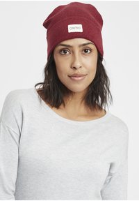 Red knit beanie hat with a label reading "oxmo." The texture is ribbed, designed to fit snugly, and has a slouchy appearance at the top.