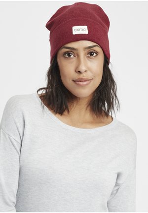 Berretto di lana a maglia rossa con un'etichetta che riporta la scritta "oxmo". La trama è a coste, progettato per adattarsi bene e presenta un aspetto scivoloso nella parte superiore.