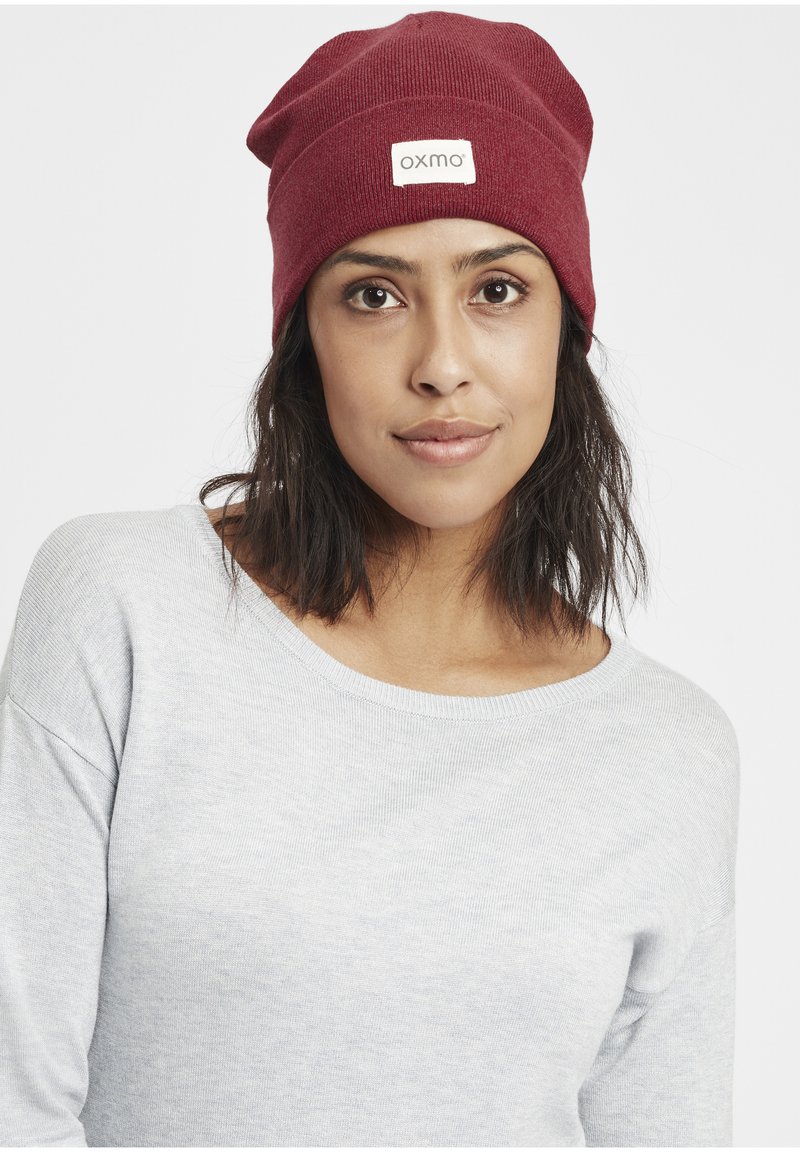 Red knit beanie hat with a label reading "oxmo." The texture is ribbed, designed to fit snugly, and has a slouchy appearance at the top.
