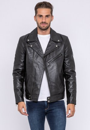 Homme portant une veste en cuir noire de motard par-dessus une chemise blanche et un jean bleu, debout devant un fond clair uni.