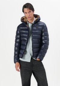 Marineblauwe pufferjack heeft een ritssluiting aan de voorkant, een capuchon en horizontale quiltstructuur. Het heeft een contrasterende beige voering en een klein logo op de borst.