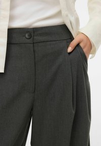 Szare, dopasowane spodnie o gładkiej fakturze, z plisami z przodu i pojedynczym czarnym guzikiem zapięcia w pasie. Odpowiednie do formalnego stroju.