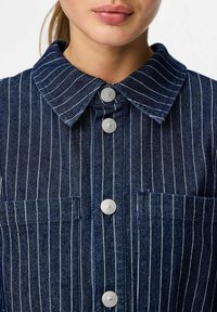 Chaqueta de mezclilla azul oscuro con rayas verticales blancas, presenta un collar puntiagudo, botones plateados y dos bolsillos frontales.