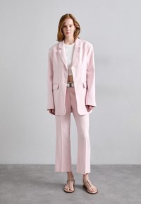 Blazer ajusté rose clair avec deux poches avant et deux boutons, associé à un pantalon évasé assorti. Top cropped blanc et sandales décontractées.