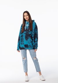 Felpa oversize teal e nera con effetto tie-dye, dotata di tasca frontale e cordini, abbinata a jeans chiari strappati e sneaker bianche.