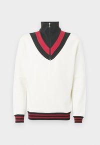 Valge pullover kõrge kaelakaelusega, millel on musta ja punase V-kujuline muster rinnal ning kontrastsed soonikkoes mansetid ja alumine ääris.