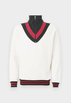 Bela majica s puloverjem z visokim ovratnikom, na prsih s črno-rdečim V-obliki in kontrastnimi rebrastimi manšetami ter robom.