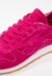 Vaaleanpunaiset mokkanahkakengät, joissa on nauhakiinnitys ja kohokuvioitu "Reebok"-logo. Varusteena valkoinen välikenk�ä ja vaalea kumipohja.