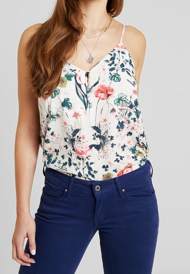 Débardeur floral en tissu léger avec des fleurs roses, vertes et bleues sur un fond crème ; associé à un pantalon bleu marine.