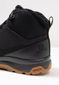 Κοντινή λήψη μαύρου ορειβατικού μποτακιού με επενδεδυμένο γιακά, ταινία έλξης και λάστιχο σόλας με την επιγραφή "contagrip", που φέρει το λογότυπο της Salomon στο πλάι.