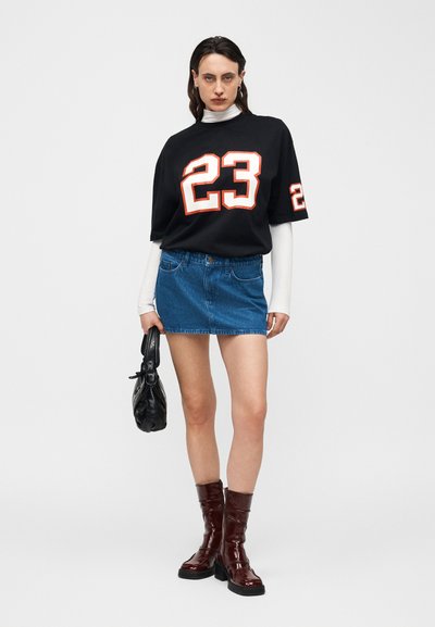 Kvinne iført svart oversized t-skjorte med nummer 23, hvit langermet topp under, blå denim miniskjørt, brune støvler, holder en svart håndveske.