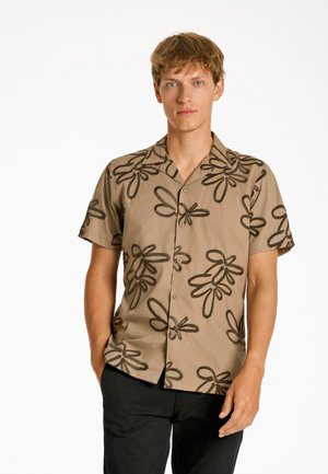 Ung mand iført en beige kortærmet skjorte med store mørke abstrakte blomsterprints, stående med den ene hånd i lommen.