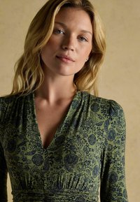 Zielona sukienka z wzorzystym wzorem w granatowym kolorze, z głębokim dekoltem w kształcie litery V, marszczonym pasem i długimi rękawami. Widoczna miękka tekstura materiału.