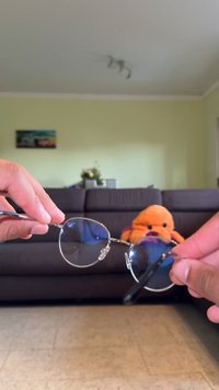 Lunettes de soleil rondes avec des verres teintés foncés, monture en métal argenté et plaquettes de nez ajustables, tenues par deux mains contre un arrière-plan flou.