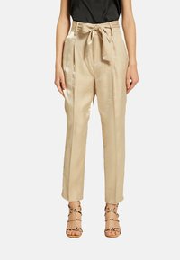 Pantalons beiges taille haute avec une ceinture à nouer, devant plissé et texture lisse et brillante. Associés à des sandales à lanières.