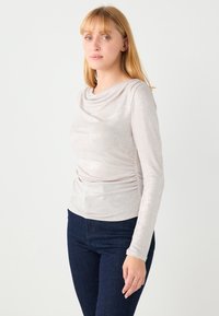 Długi rękaw w jasnym metalicznym beżu z luźnym dekoltem, ozdobiony marszczeniem z boku, zestawiony z ciemnoniebieskimi dżinsami.