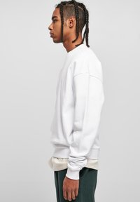 Hvid crewneck sweatshirt lavet af blødt stof, med lange ærmer og ribbede manchetter. Båret over et lys kontrastlag, set fra siden.