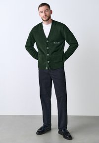Green knitted cardigan with a v-neckline, button front, two front pockets, and ribbed hem. Paired with dark pants and black shoes.
