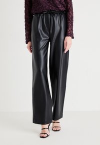 Pantalons larges en simili cuir noir avec une taille élastique et un cordon de serrage. Texture lisse et coupe ajustée, associés à des sandales noires.