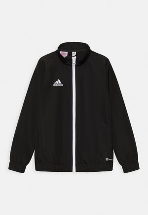 Giacca da track nera Adidas con colletto alto, zip frontale completa, logo bianco e polsini e orlo elasticizzati. Due tasche laterali visibili.