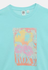 Chemise bleu clair avec des fleurs d'hibiscus roses, des palmiers, un soleil jaune, et le texte "ROXY" en couleurs pastel sur le devant.