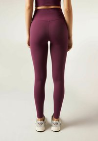 Vysoké, elastické legíny z hlubokého burgundského materiálu. Hladký povrch s přiléhavým střihem, doplněné o šedé sportovní boty.