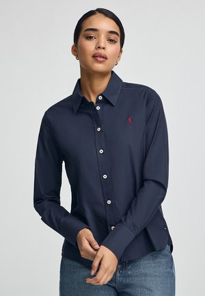 Chemise boutonnée bleu marine en tissu lisse avec des manches longues, des boutons blancs et un petit logo brodé rouge sur la poitrine gauche.
