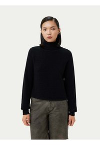 Svart ribbad polotröja med långa ärmar, med en texturerad stickad design. Kombinationen av grå byxor, på en neutral bakgrund.