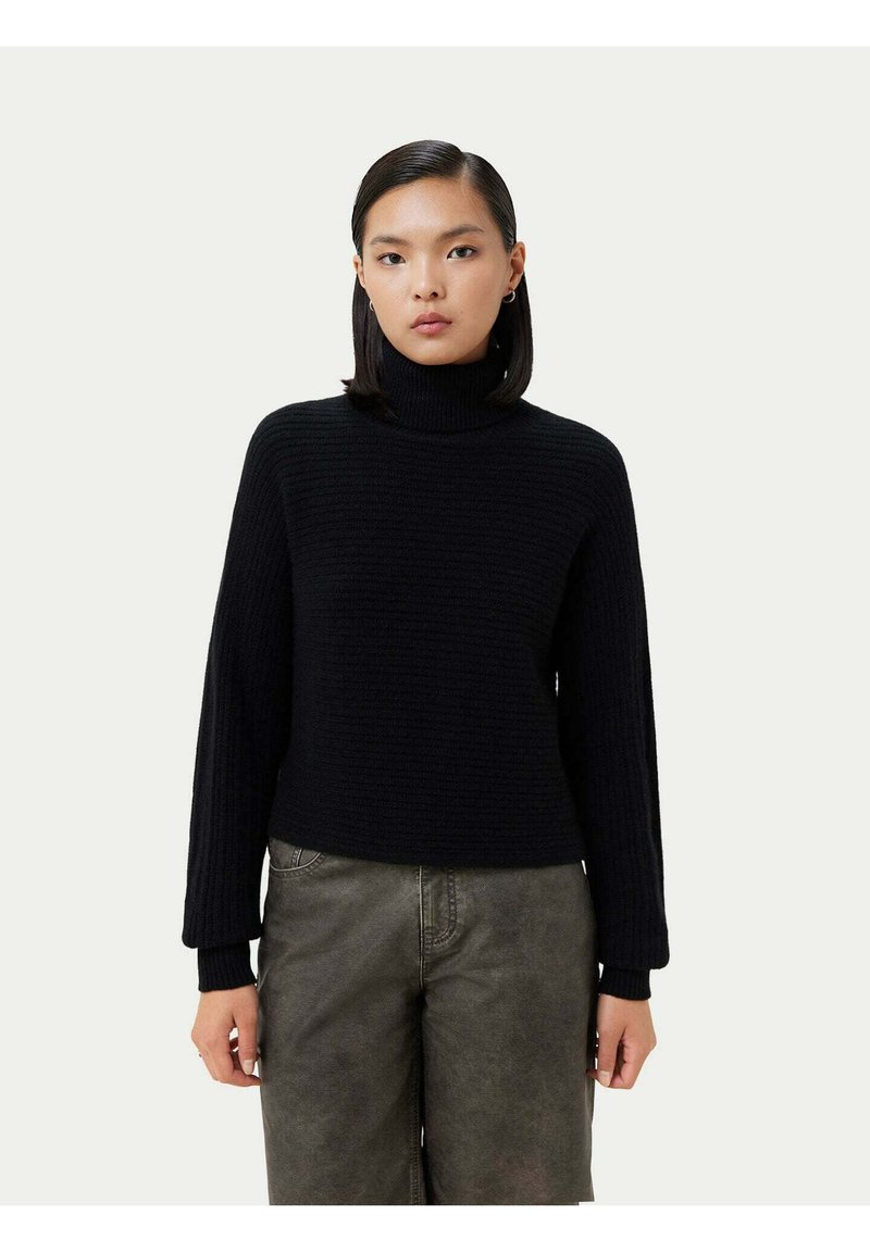 Svart ribbad polotröja med långa ärmar, med en texturerad stickad design. Kombinationen av grå byxor, på en neutral bakgrund.