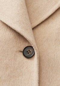 Beige wollen jas met een brede revers en een grote donkerbruine viergaatjesknop aan de voorkant.