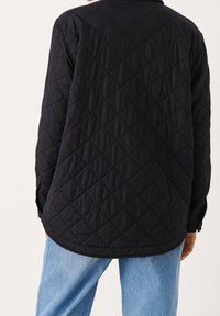 Svart vadderad jacka med diamantmönster, långa ärmar och en avslappnad passform. Har en krage och är kombinerad med ljusblå jeans.