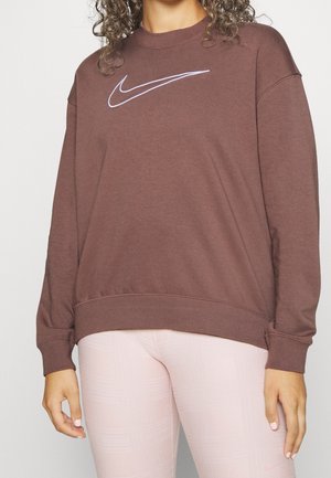 Personne portant un sweat-shirt Nike marron avec un logo blanc et des leggings rose clair, debout devant un fond blanc uni.