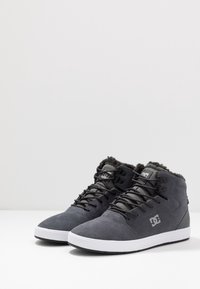 Tossut, joissa on korkea vartalo, tummanharmaata mokkaa ja valkoinen kumipohja. Ominaisuuksiin kuuluu harmaat tekstiilivälikkeet ja mustat nauhat. Logo sivulla.